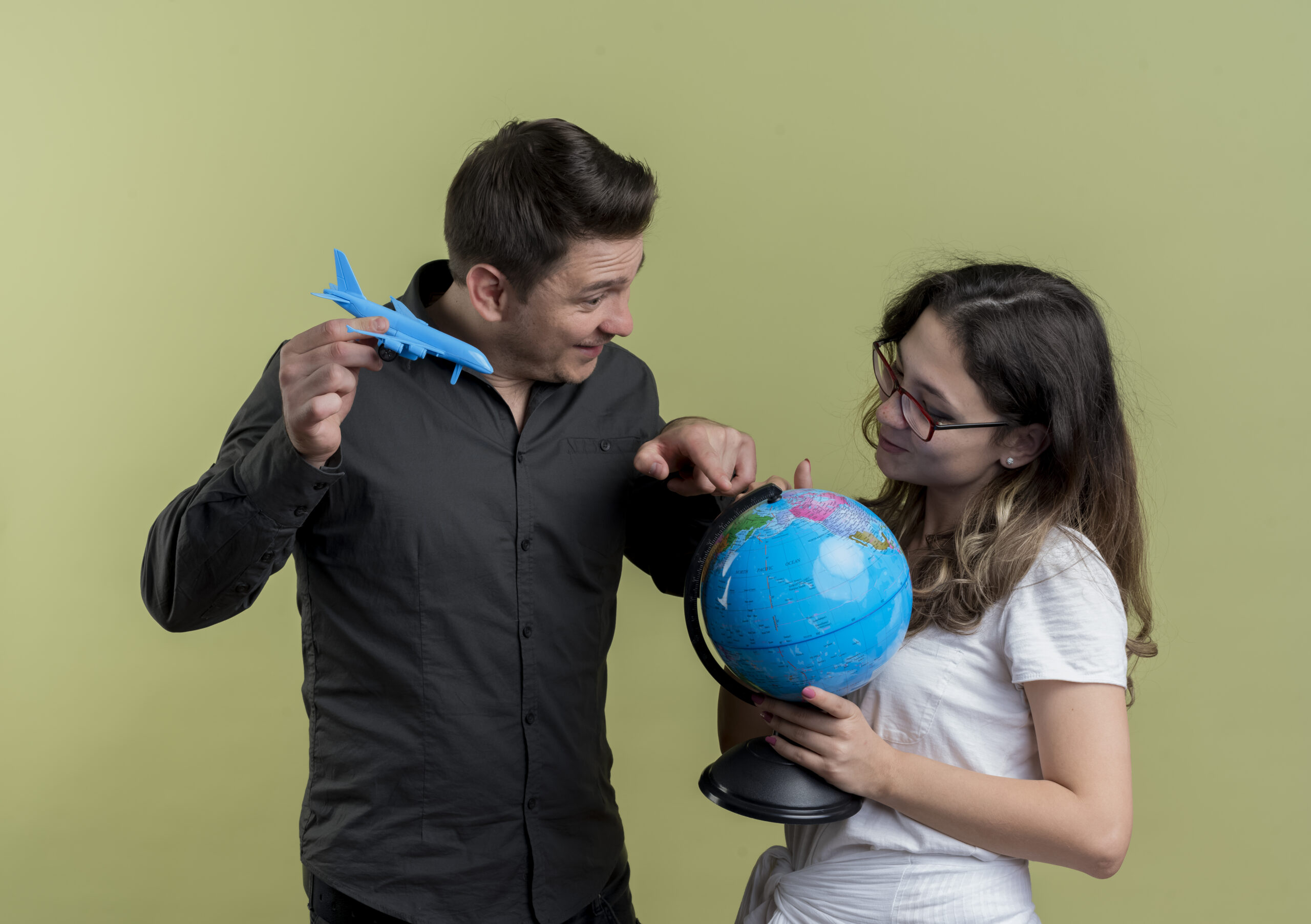 happy young couple of tourists man and woman holding globe and toy air plane standing together having fun over light background